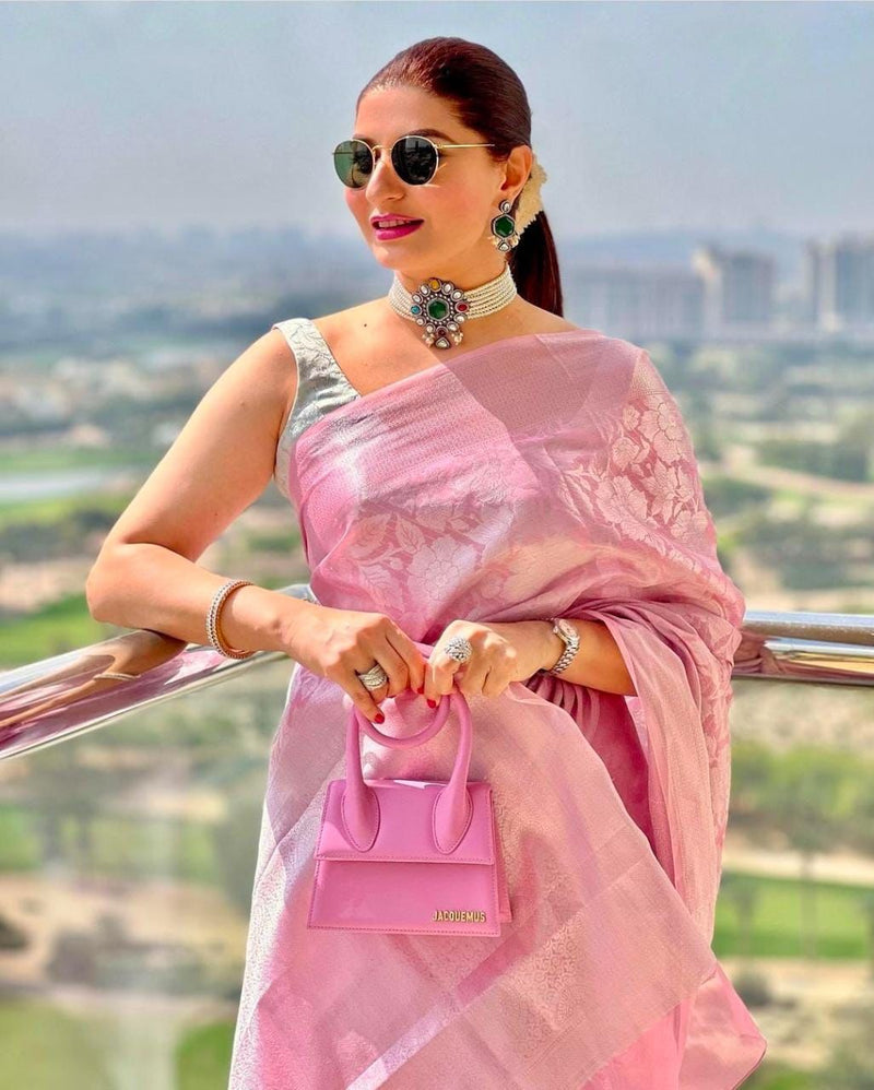 Woman in a pink saree with sunglasses and a pink handbag, standing outdoors with a scenic background.