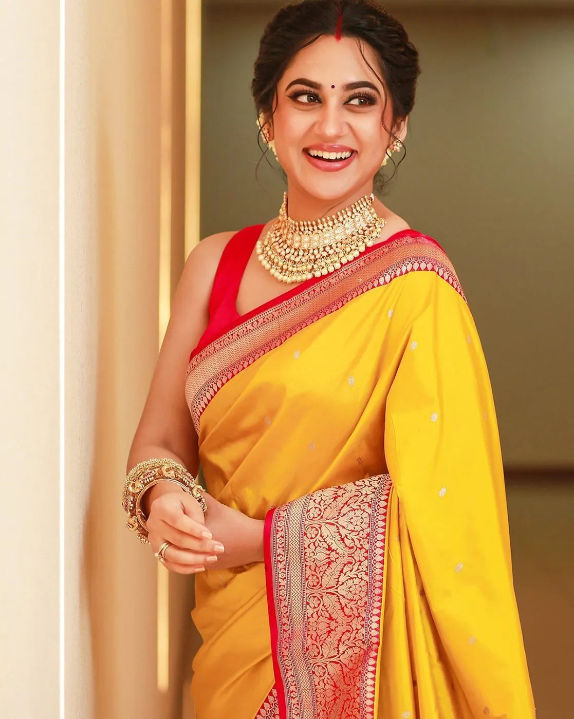 Woman in a yellow saree with red blouse and gold jewelry, smiling.