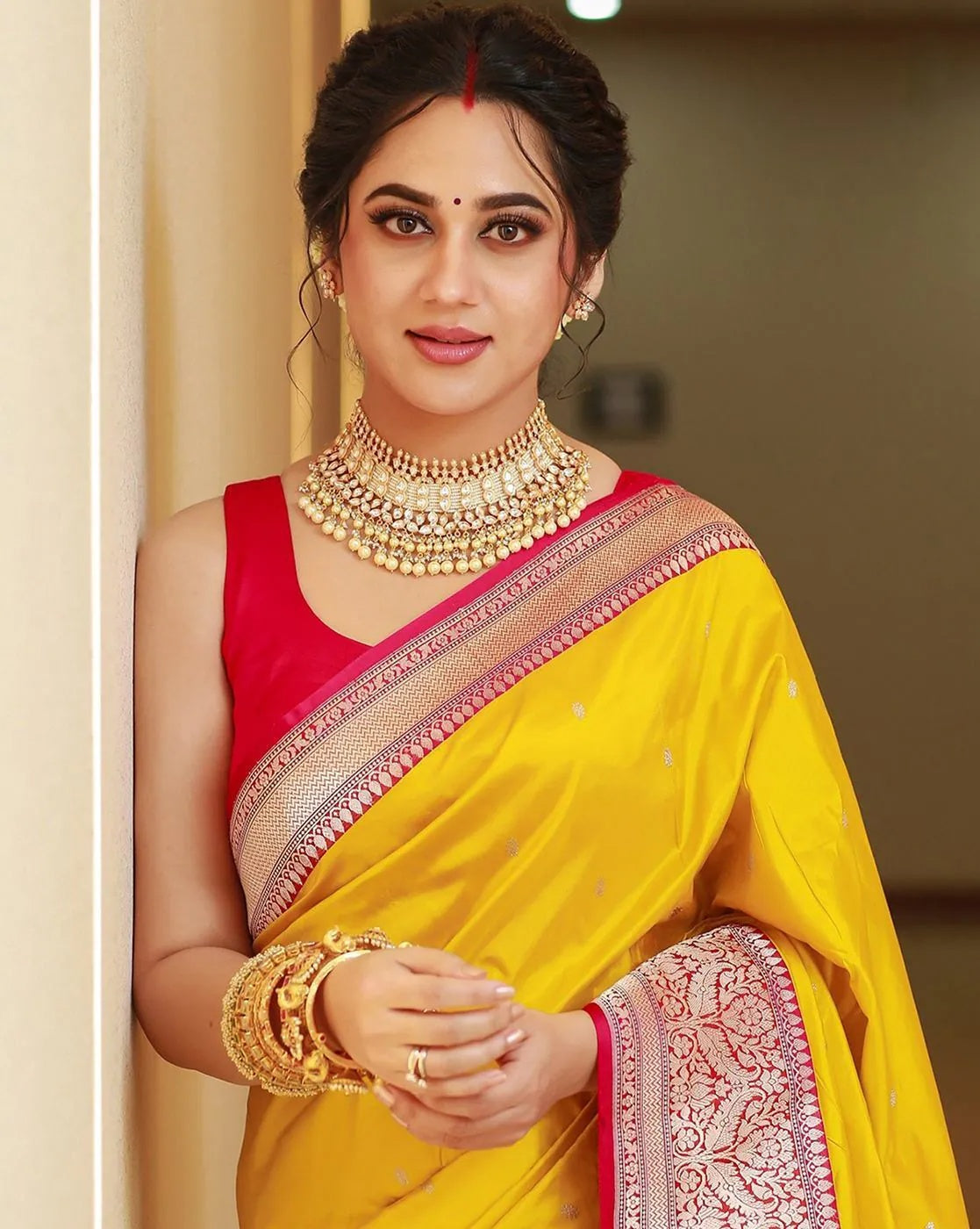 Woman in a yellow saree with red blouse, wearing gold jewelry indoors.