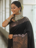 Woman wearing a black saree with intricate patterns and heavy jewelry indoors.