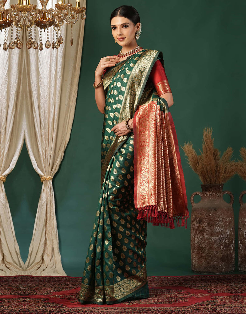 Woman in a green saree with gold patterns and a red border standing in a decorated room.