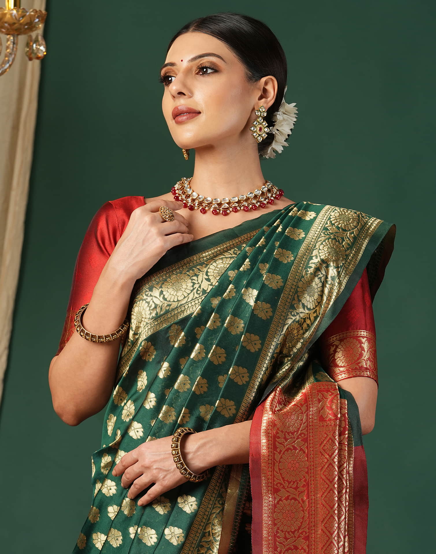 Woman wearing a green and red saree with gold jewelry against a dark green background