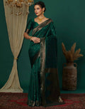 Woman in a green saree with gold border standing against a dark green wall.
