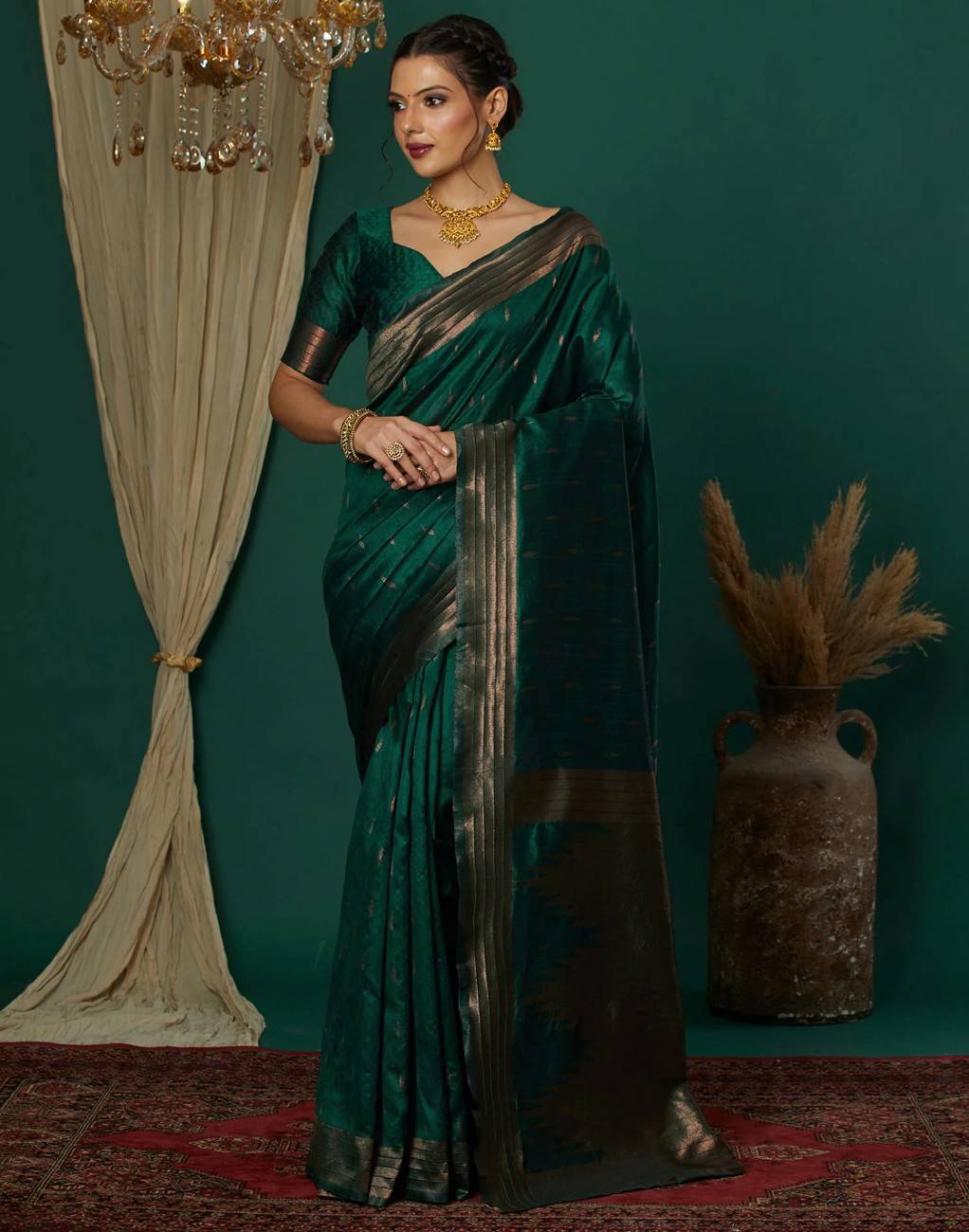Woman in a green saree with gold border standing against a dark green wall.