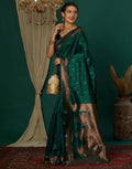 Woman in a green saree with gold details standing against a dark green wall.