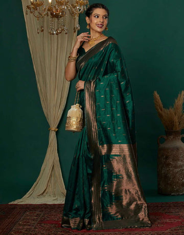 Woman in a green saree with gold details standing against a dark green wall.