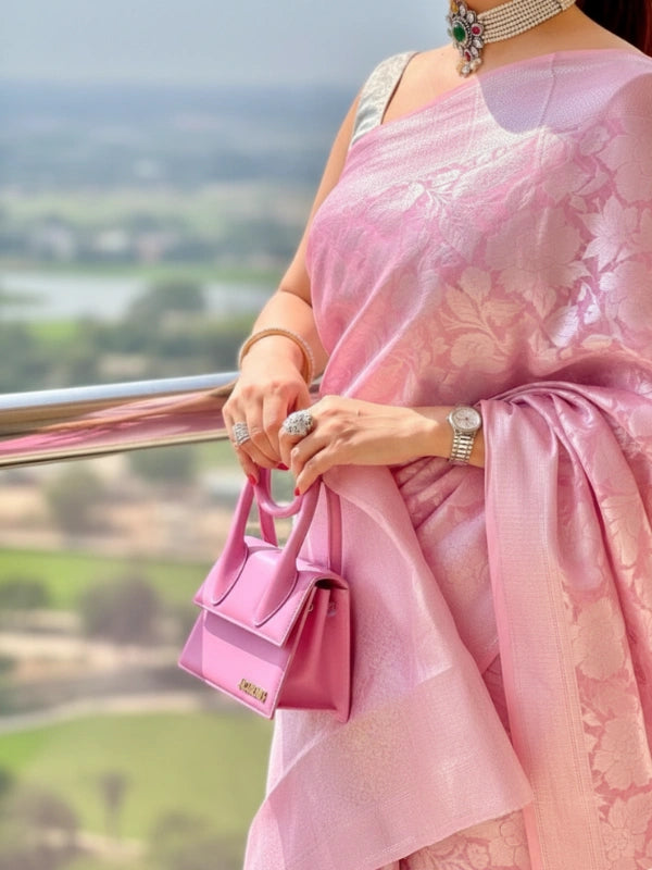 Woman in a pink saree with sunglasses and a pink handbag, standing outdoors with a scenic background.
