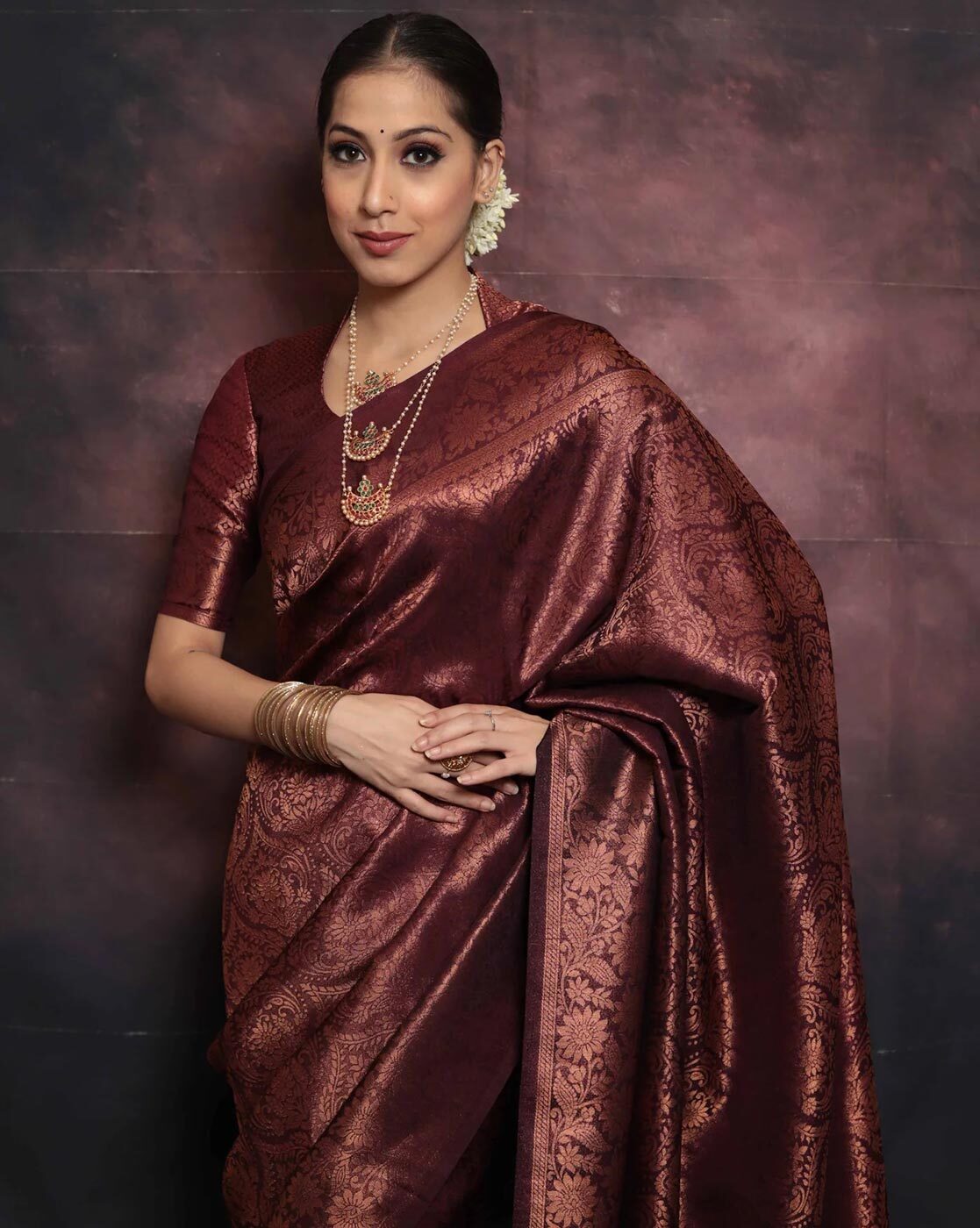 Woman wearing a maroon saree with gold patterns against a dark background