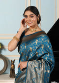 Woman wearing a blue saree with a pattern, smiling and adjusting her hair against a light blue wall.