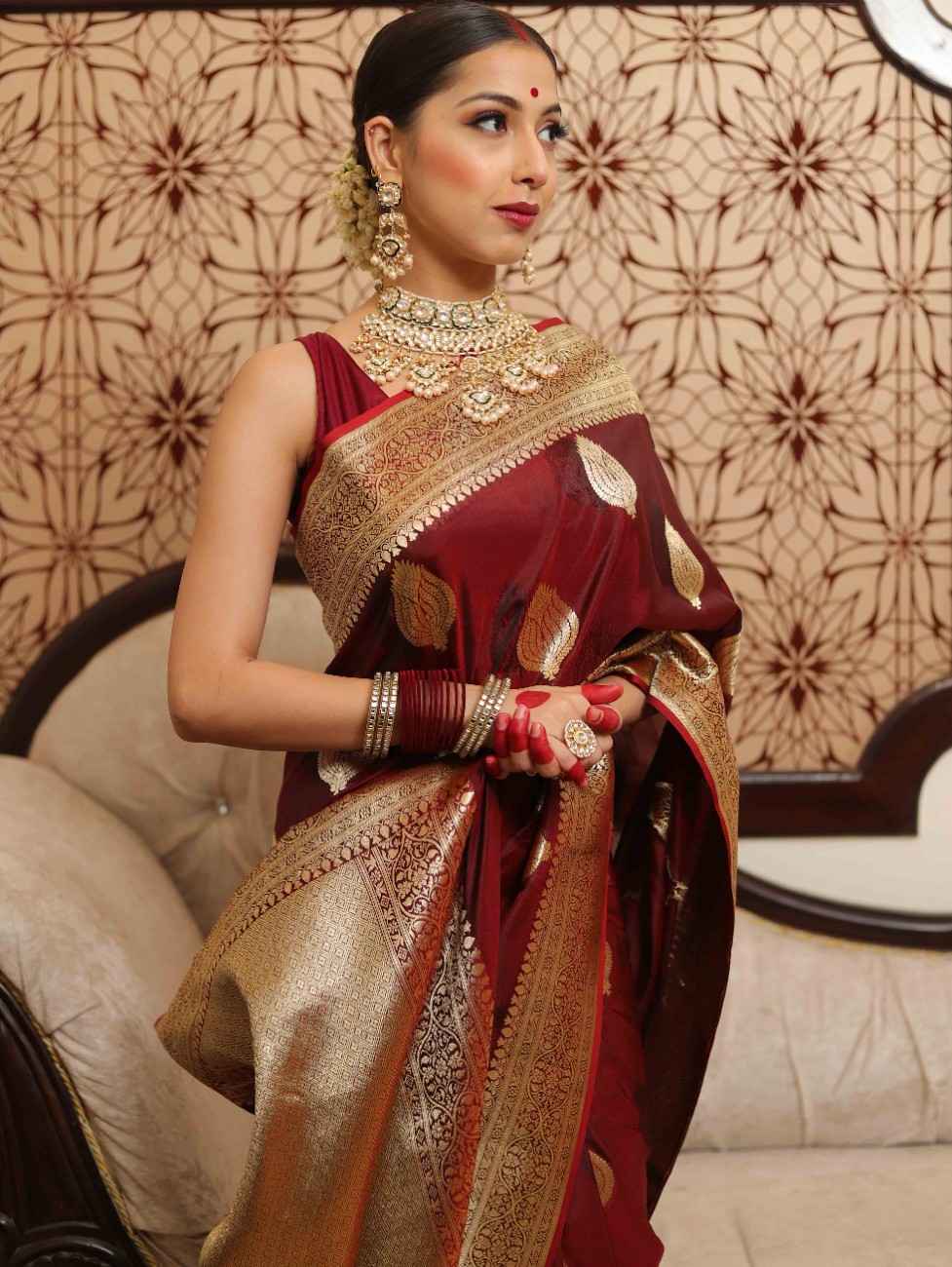 Woman in a red and gold saree with jewelry against a patterned wall.