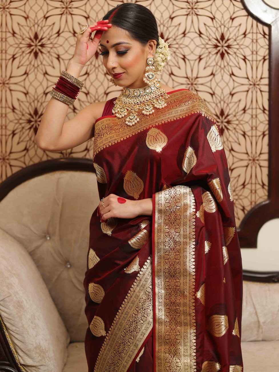 Woman in a maroon saree with gold patterns sitting on a couch.