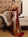 Woman in a maroon saree with gold patterns standing in a room with a decorative wall and sofa.