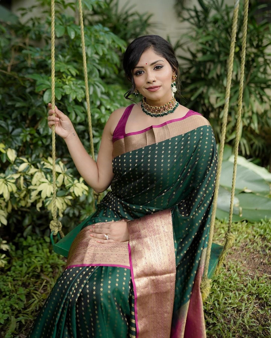 Woman in a green saree with gold patterns sitting on a swing outdoors.