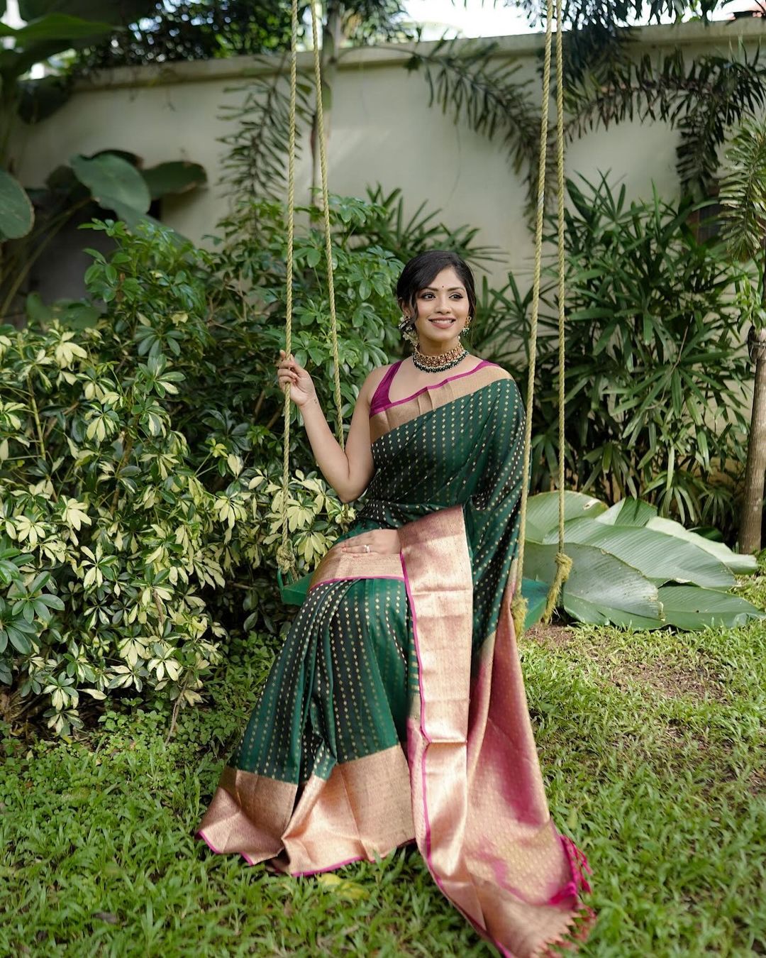 Woman in a green and pink saree sitting on a swing with greenery in the background
