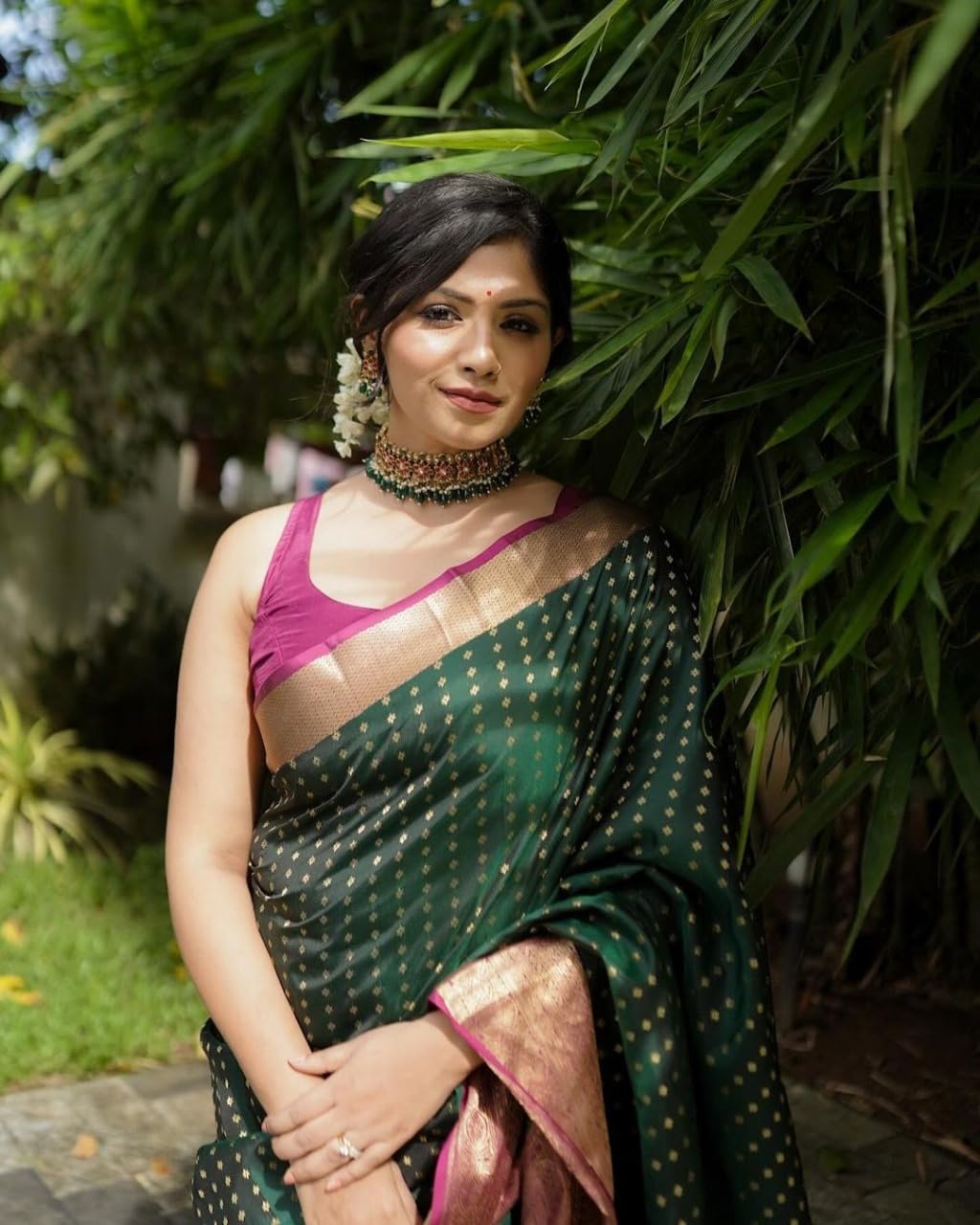 Woman in a green saree with a pink blouse standing outdoors among greenery