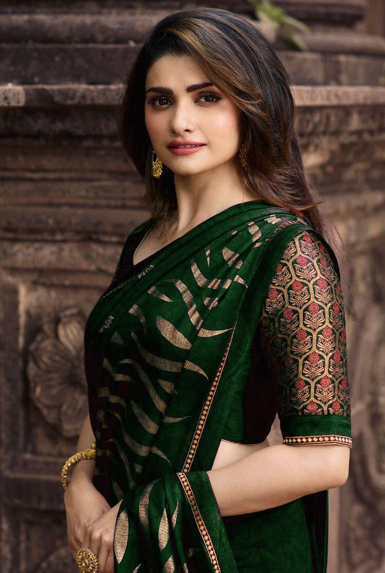 Woman in a green saree with black patterns standing in front of stone architectural elements.